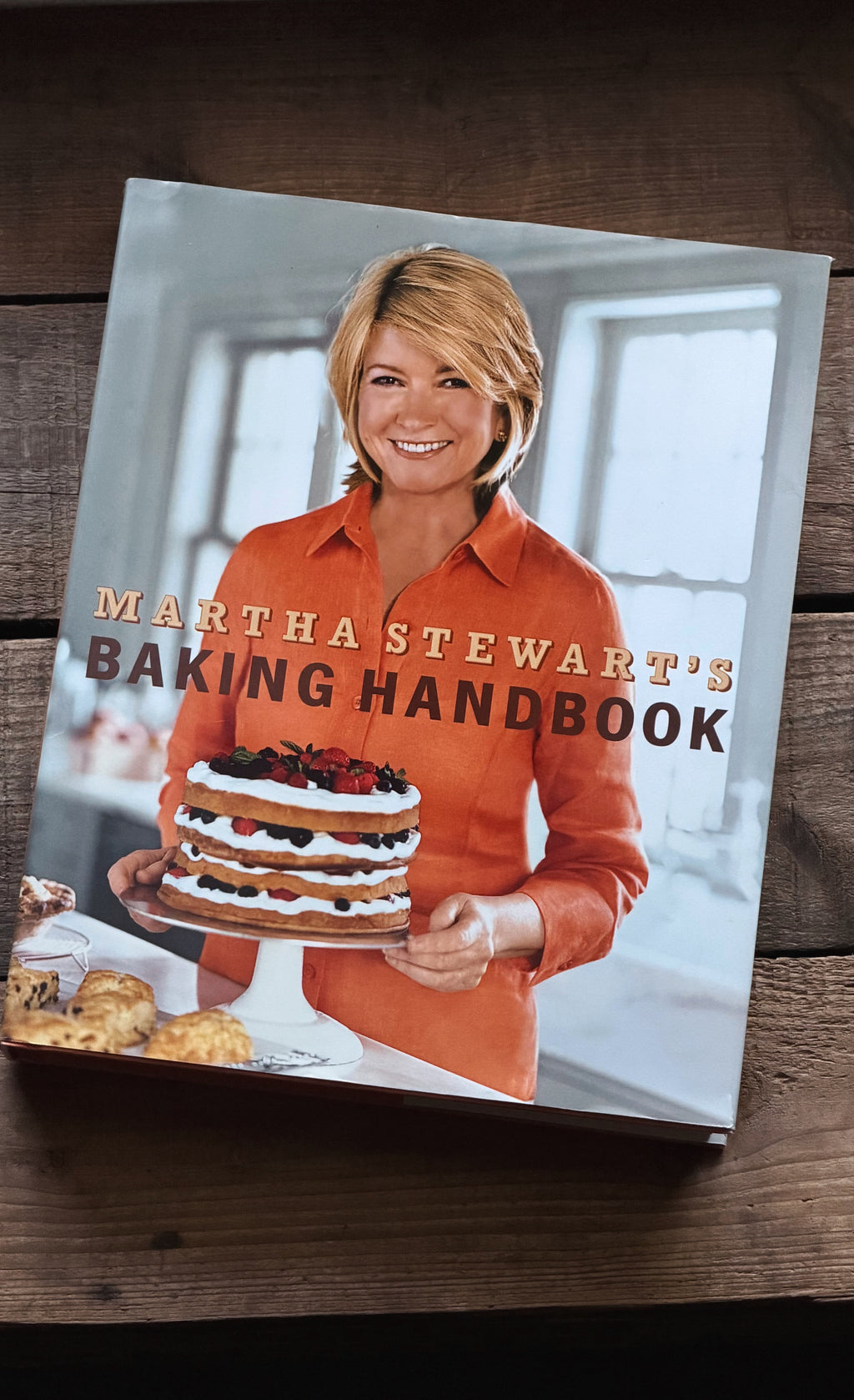 The front cover of the book 'Martha Stewart's Baking Handbook' is displayed on a dark wooden surface. The cover features a photo of Martha Stewart holding a multi-layered cake, wearing a bright orange shirt.