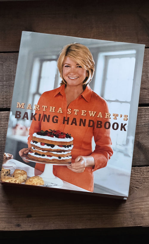 The front cover of the book 'Martha Stewart's Baking Handbook' is displayed on a dark wooden surface. The cover features a photo of Martha Stewart holding a multi-layered cake, wearing a bright orange shirt.
