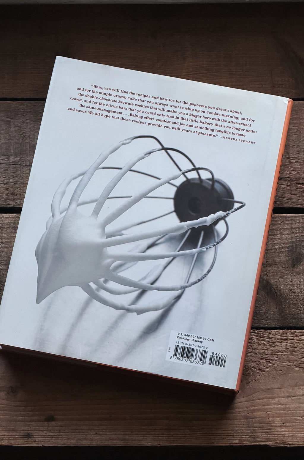 The back cover of a book resting on a dark wooden surface. The cover features a black and white photograph of a hand holding a white wire whisk with a black ball, with a small orange spine edge visible on the right.