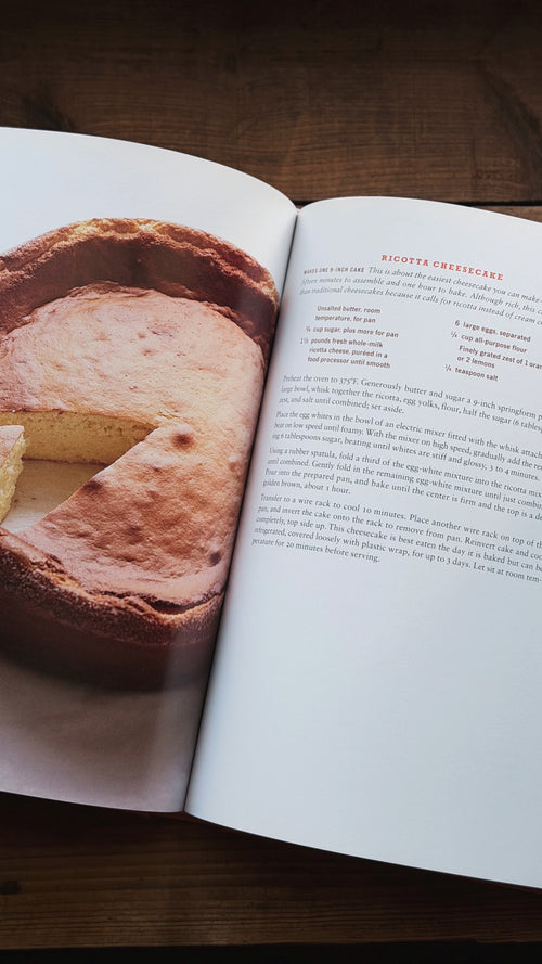 An open cookbook spread on a dark wooden table, showing a photo of a cheesecake on the left page and recipe text on the right page.