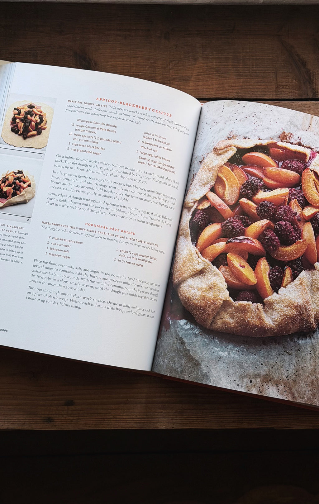 An open cookbook spread on a dark wooden table, showing a recipe page with text and photos on the left and a large photo of a berry and peach galette on the right.