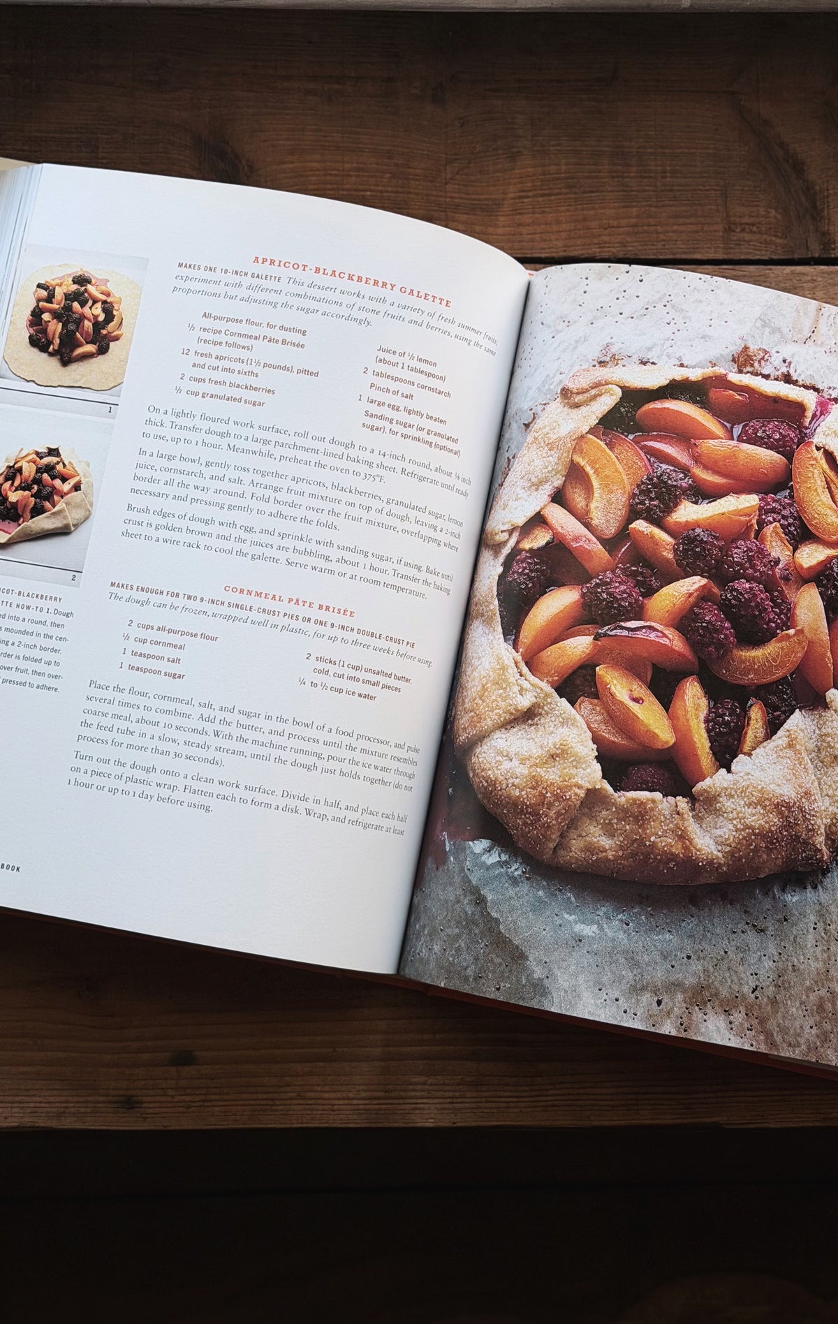 An open cookbook spread on a dark wooden table, showing a recipe page with text and photos on the left and a large photo of a berry and peach galette on the right.