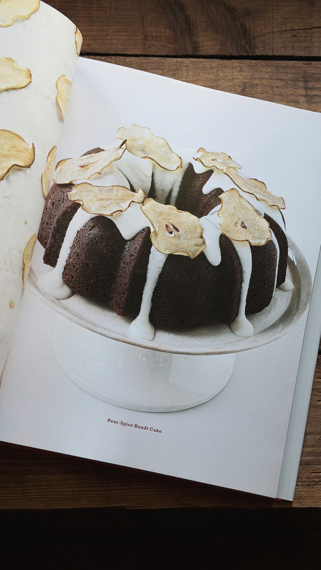 An open cookbook or recipe book on a dark wooden table, showing a printed photograph of a chocolate ring cake with white icing and dried apple slices on a white cake stand.