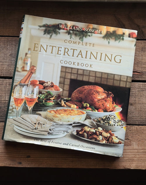Williams-Sonoma Complete Entertaining Cookbook resting on a dark wooden surface. The book cover features a festive holiday spread with a roasted turkey, plates, champagne, and candles.