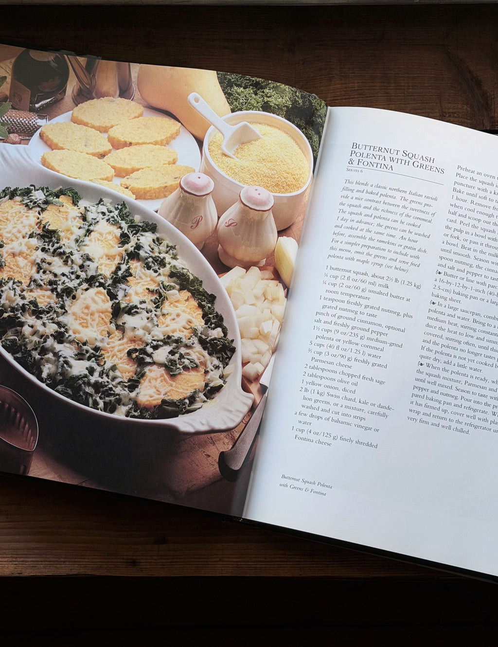An open cookbook spread on a dark wooden table, showing a large color photograph of a butternut squash casserole on the left page and printed recipe text on the right page.