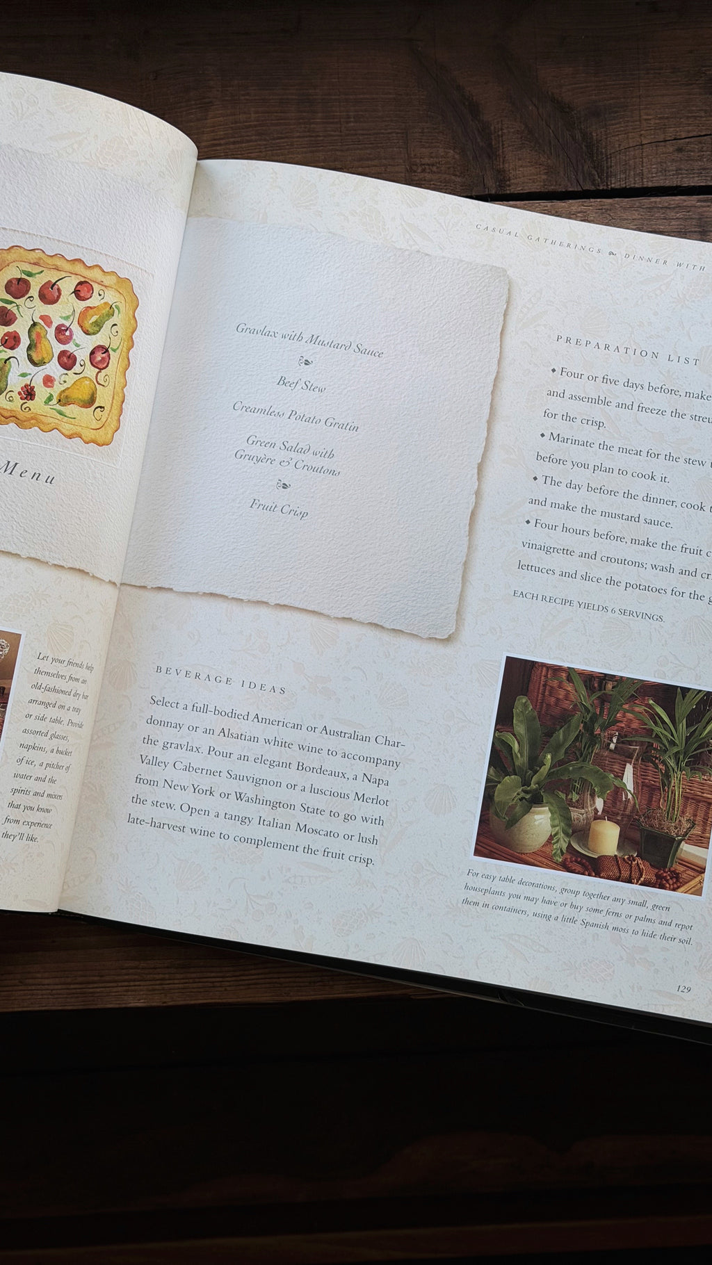 An open printed recipe book spread on a dark wooden table, showing page layouts with illustrations, text, and a small food photography inset.