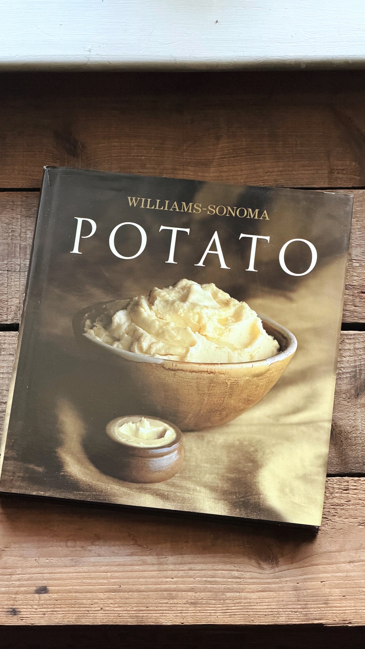 Williams-Sonoma Potato cookbook resting on a rustic wooden surface, showing the front cover with a photo of mashed potatoes in a bowl.