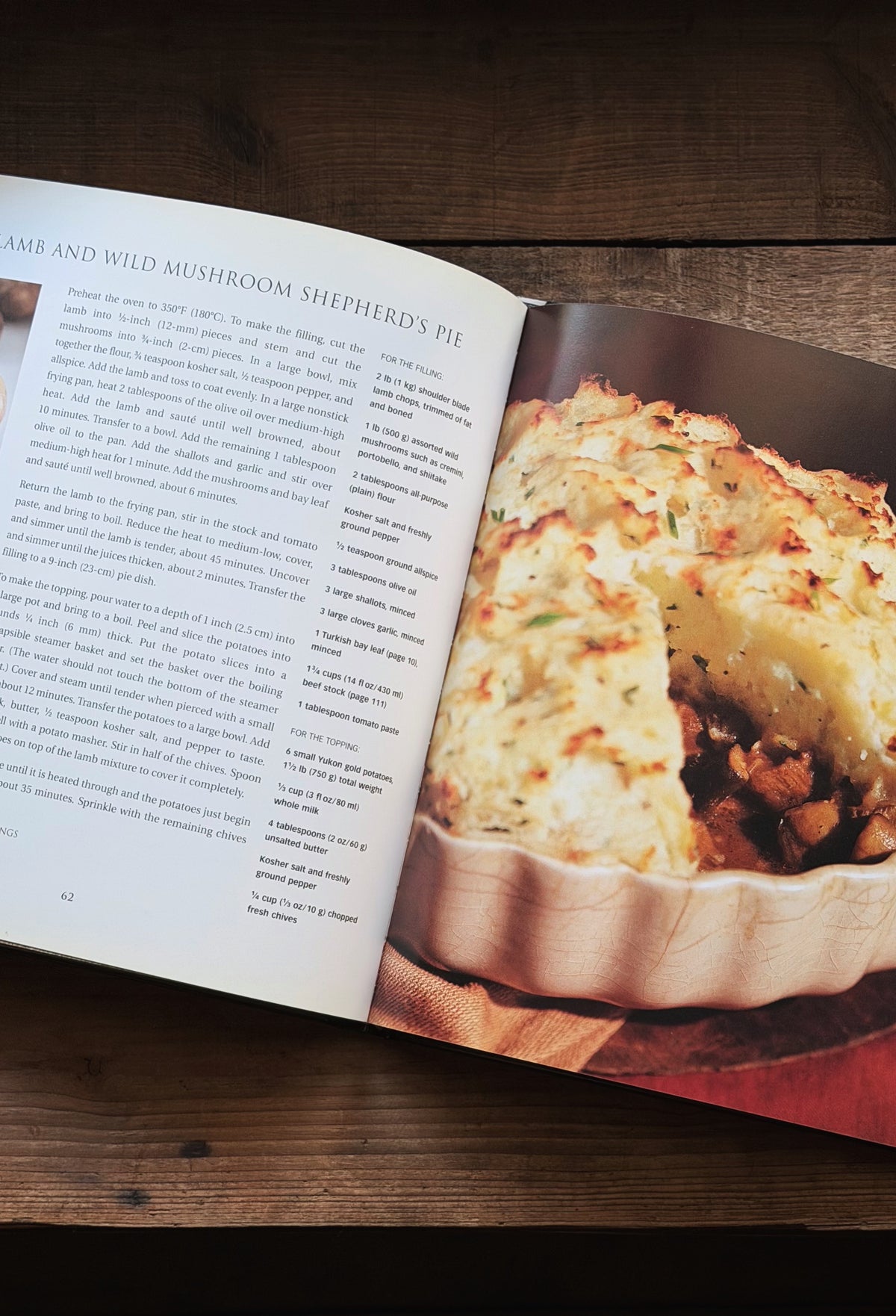 An open cookbook spread on a dark wooden table, showing a recipe page with text on the left and a photo of baked shepherd's pie on the right.