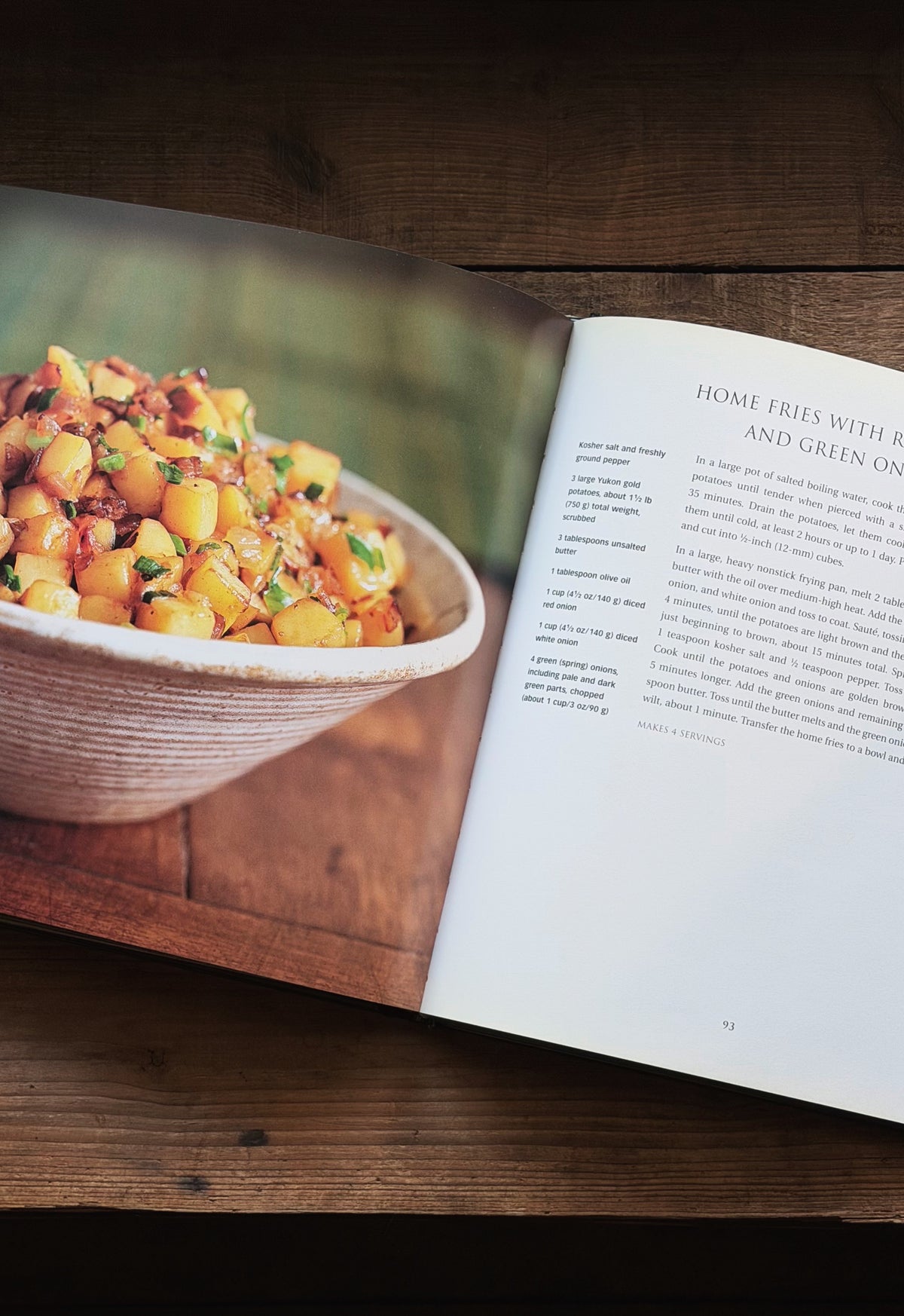 An open cookbook lying on a dark wooden table, showing a large food photograph of a bowl of fried potato dish on the left page and recipe text on the right page.