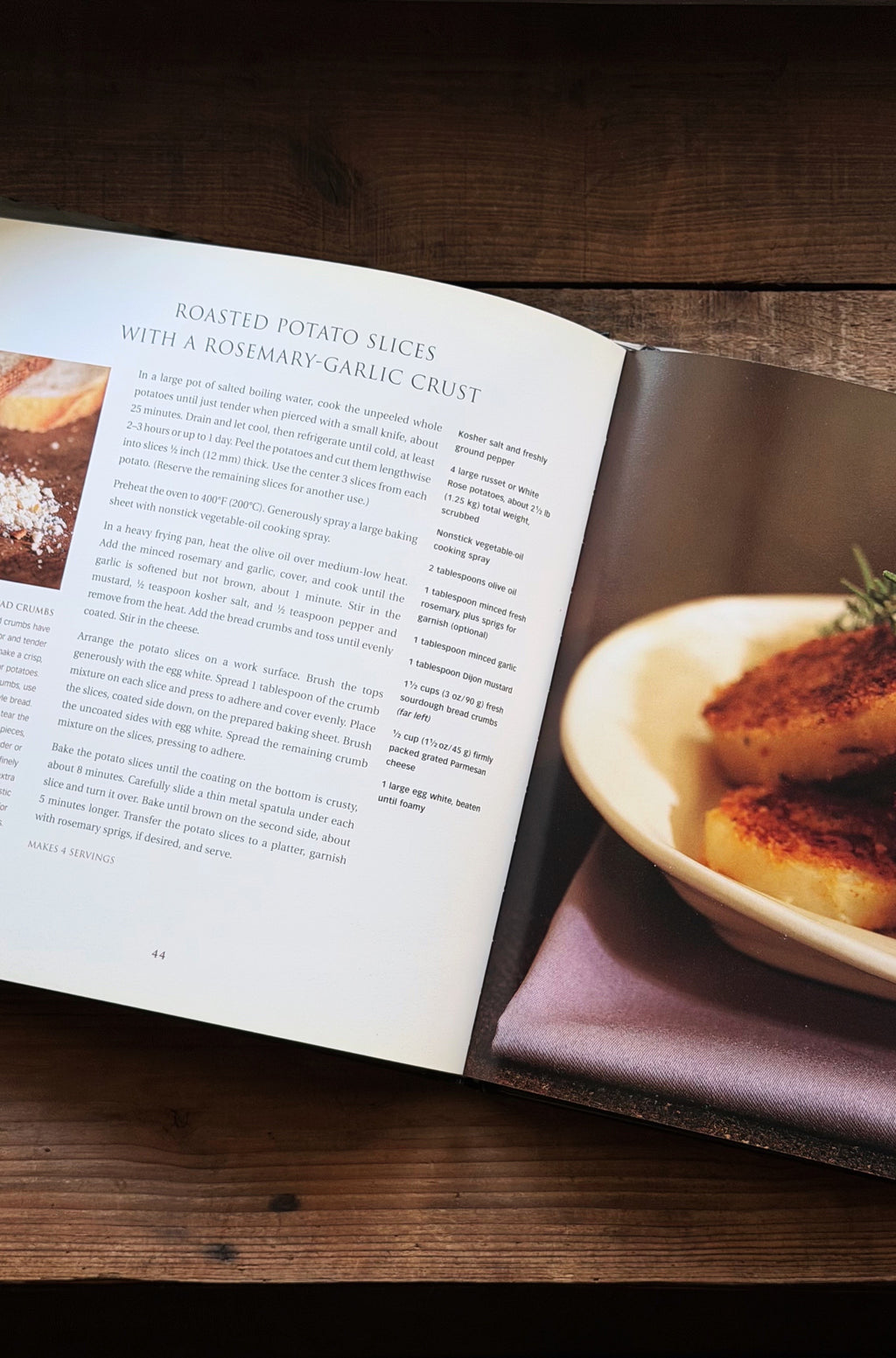 An open cookbook spread on a dark wooden table, showing a recipe page titled 'Roasted Potato Slices with a Rosemary-Garlic Crust' alongside a plated potato dish in the photo layout.