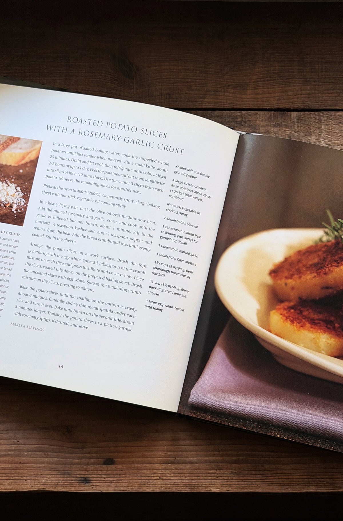 An open cookbook spread on a dark wooden table, showing a recipe page titled 'Roasted Potato Slices with a Rosemary-Garlic Crust' alongside a plated potato dish in the photo layout.
