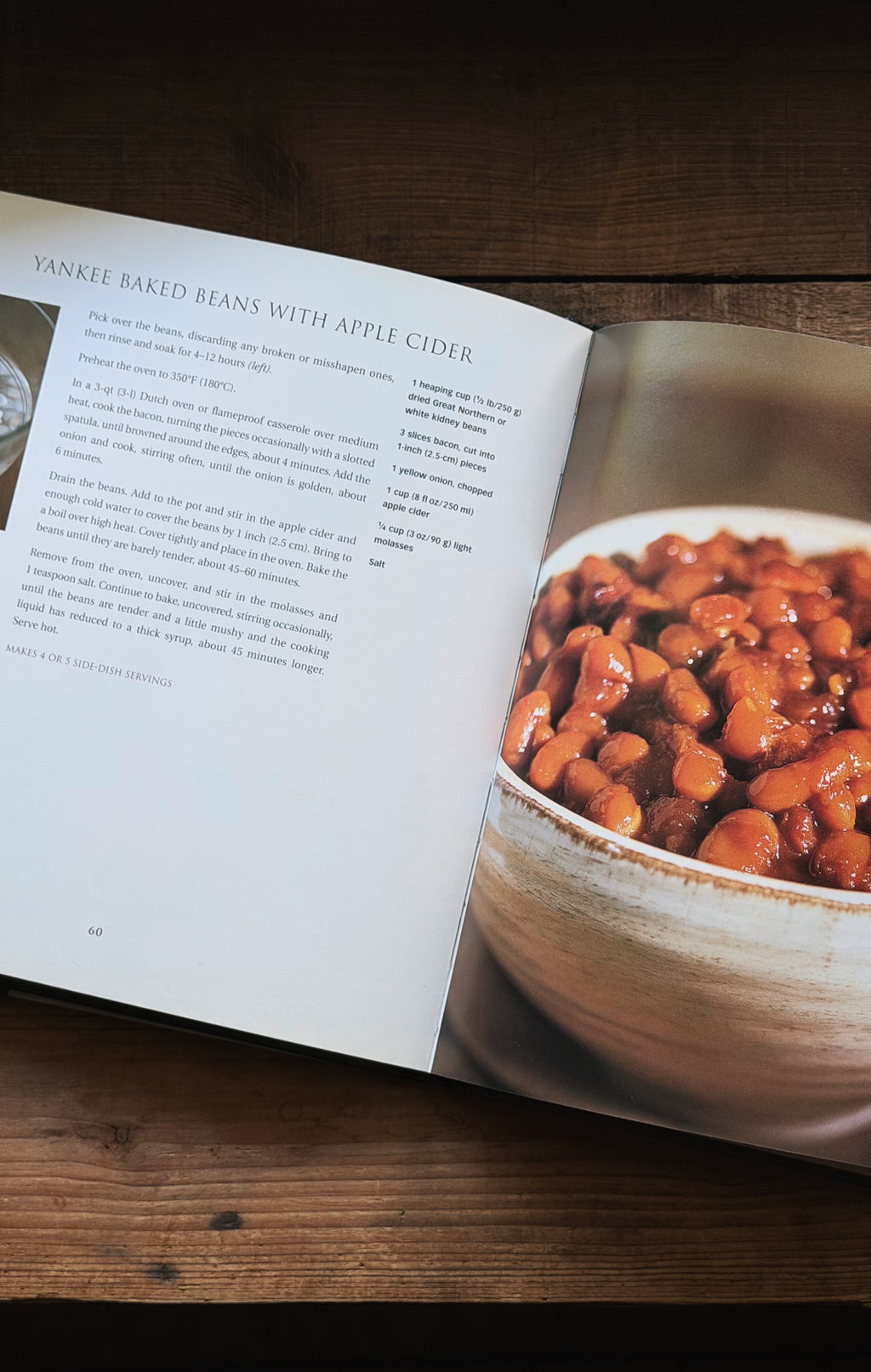An open printed cookbook or recipe book lying on a dark wooden surface, showing a page titled 'Yankee Baked Beans with Apple Cider' with text on the left page and a large photo of baked beans in a bowl on the right page.