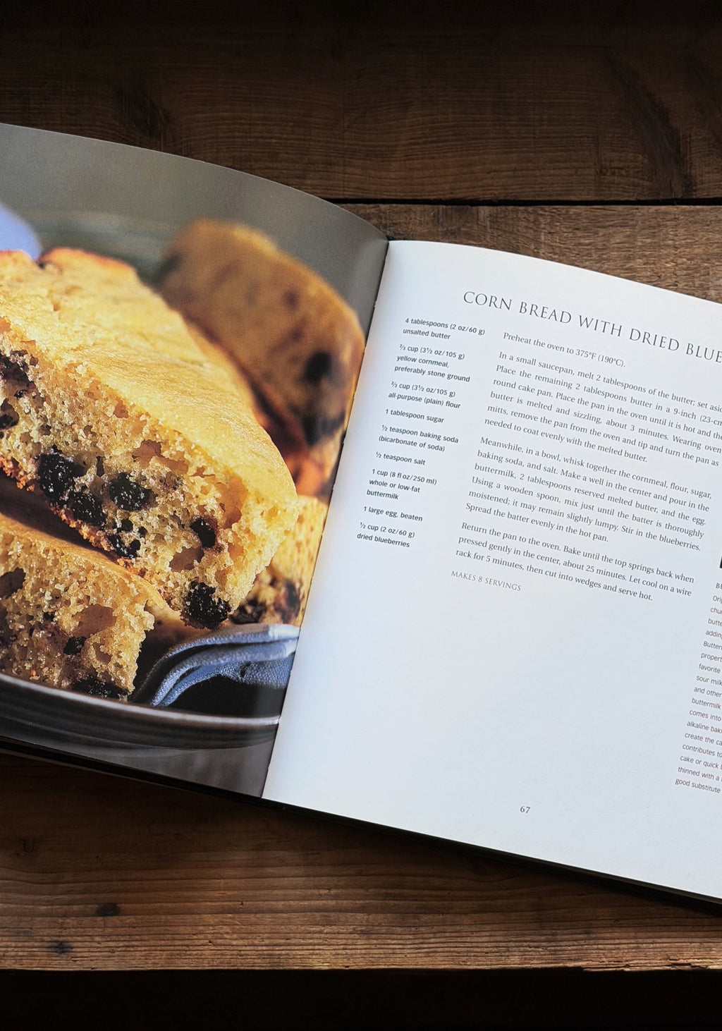 An open cookbook lying on a wooden table, showing a food photograph of corn bread with dried blueberries on one page and recipe text on the other.