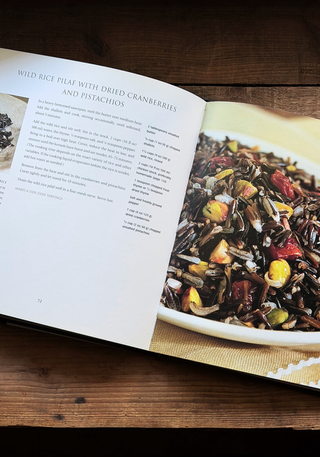 An open cookbook spread on a dark wooden table, showing a recipe page for wild rice pilaf with dried cranberries and pistachios alongside a close-up food photograph of the prepared dish on a white plate.