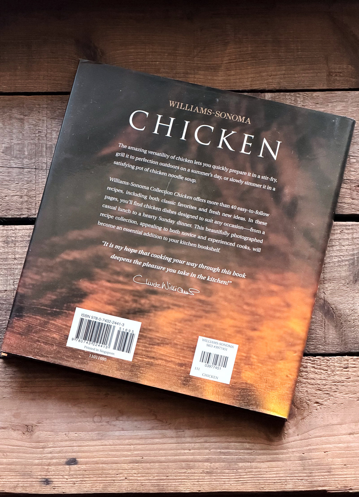 The back cover of the book 'Williams Sonoma Chicken' is displayed diagonally on a rustic wooden surface. The cover features a dramatic orange and black sunset sky over water, with the title 'CHICKEN' in large white letters and the Williams Sonoma brand name at the top.