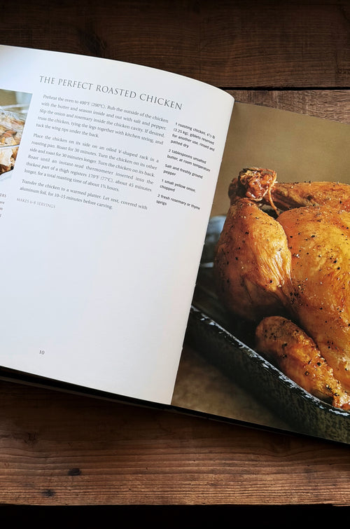 An open cookbook spread on a dark wooden table, showing the recipe page for 'The Perfect Roasted Chicken' next to a real roasted chicken in a baking dish.
