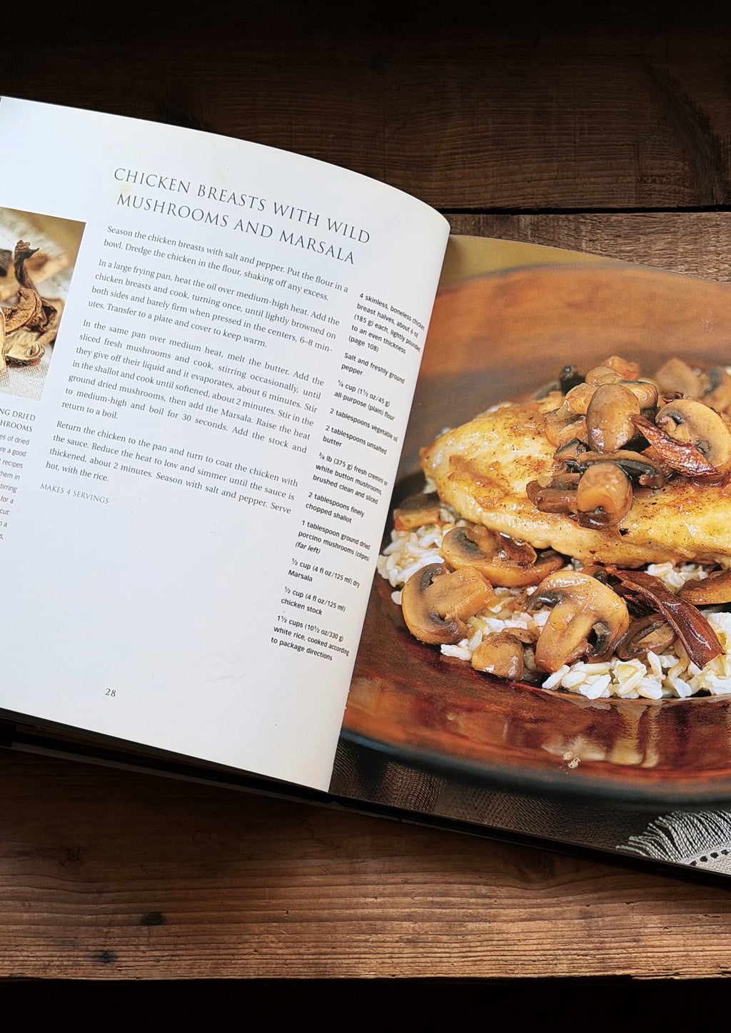 An open cookbook spread on a dark wooden table, showing a recipe page on the left and a plated dish of chicken with wild mushrooms and marsala on the right.