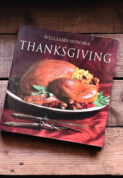 Williams-Sonoma Thanksgiving cookbook resting on a rustic wooden surface, showing the front cover with a roasted turkey and holiday garnishes.