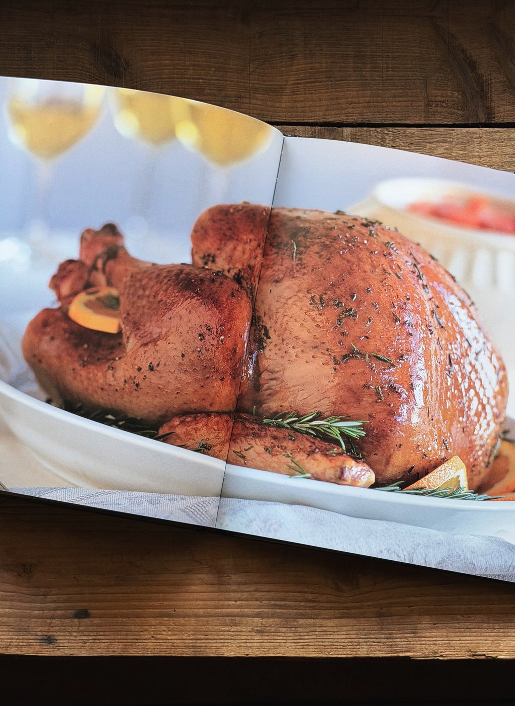 A printed cookbook or recipe book open to a page featuring a roasted turkey on a white platter, placed on a rustic wooden table.