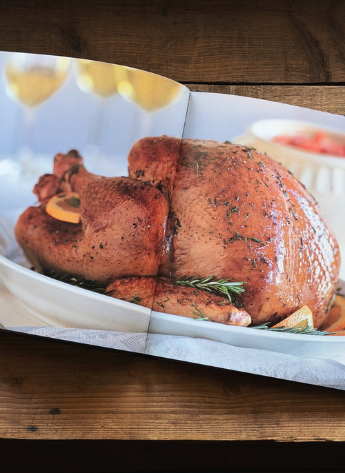 A printed cookbook or recipe book open to a page featuring a roasted turkey on a white platter, placed on a rustic wooden table.