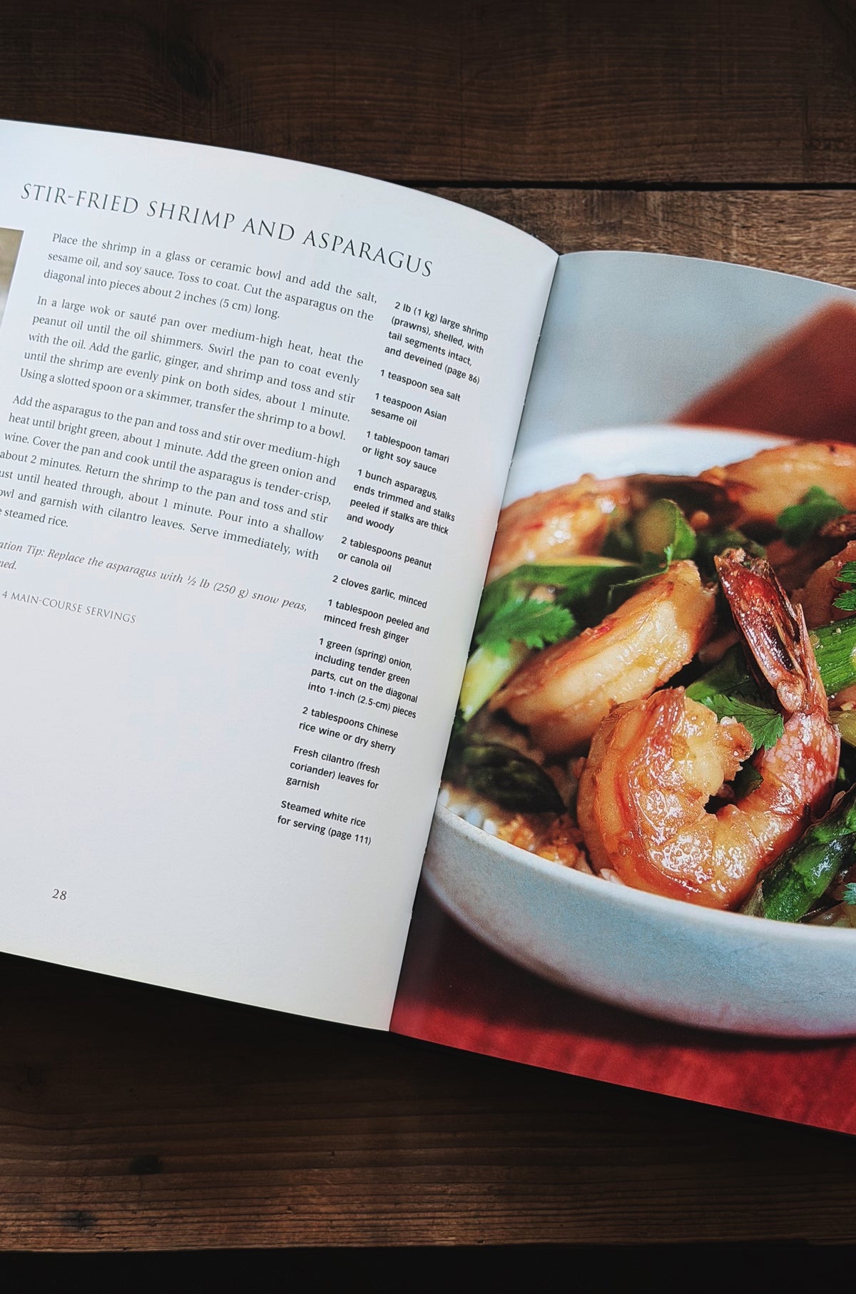An open cookbook spread showing the recipe for stir-fried shrimp and asparagus, with a photograph of the prepared dish on the right page, placed on a dark wooden surface.