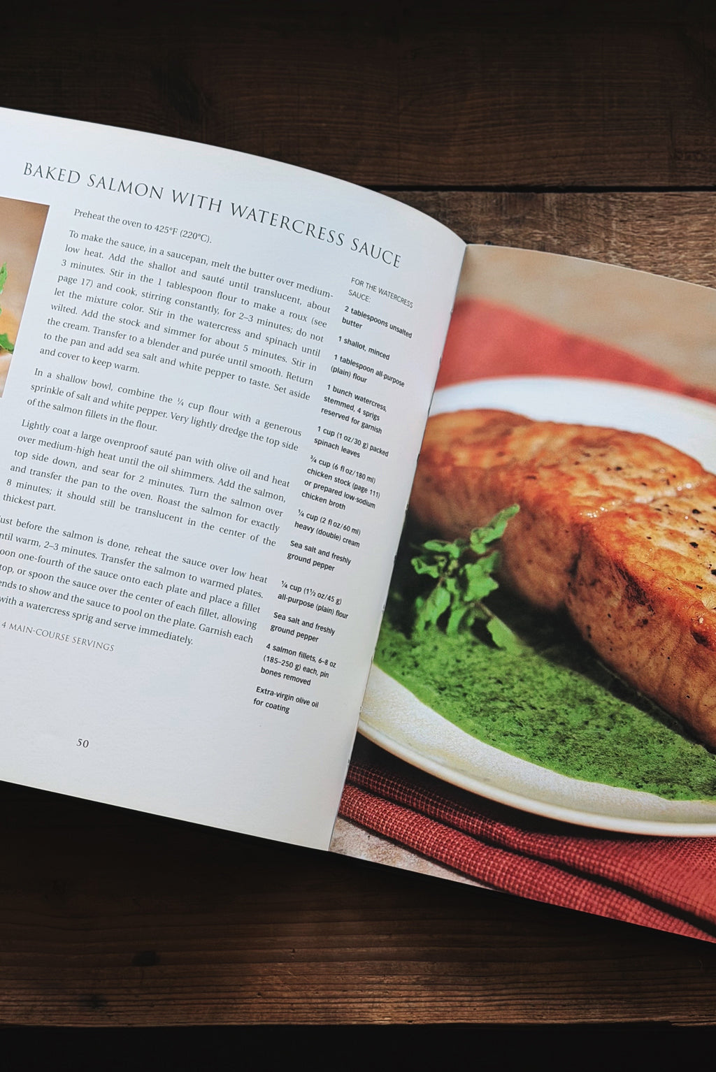 An open cookbook or recipe magazine laid on a dark wooden table, showing a page with text and a large food photograph of baked salmon with watercrress sauce on a white plate with green puree and a red cloth underneath.