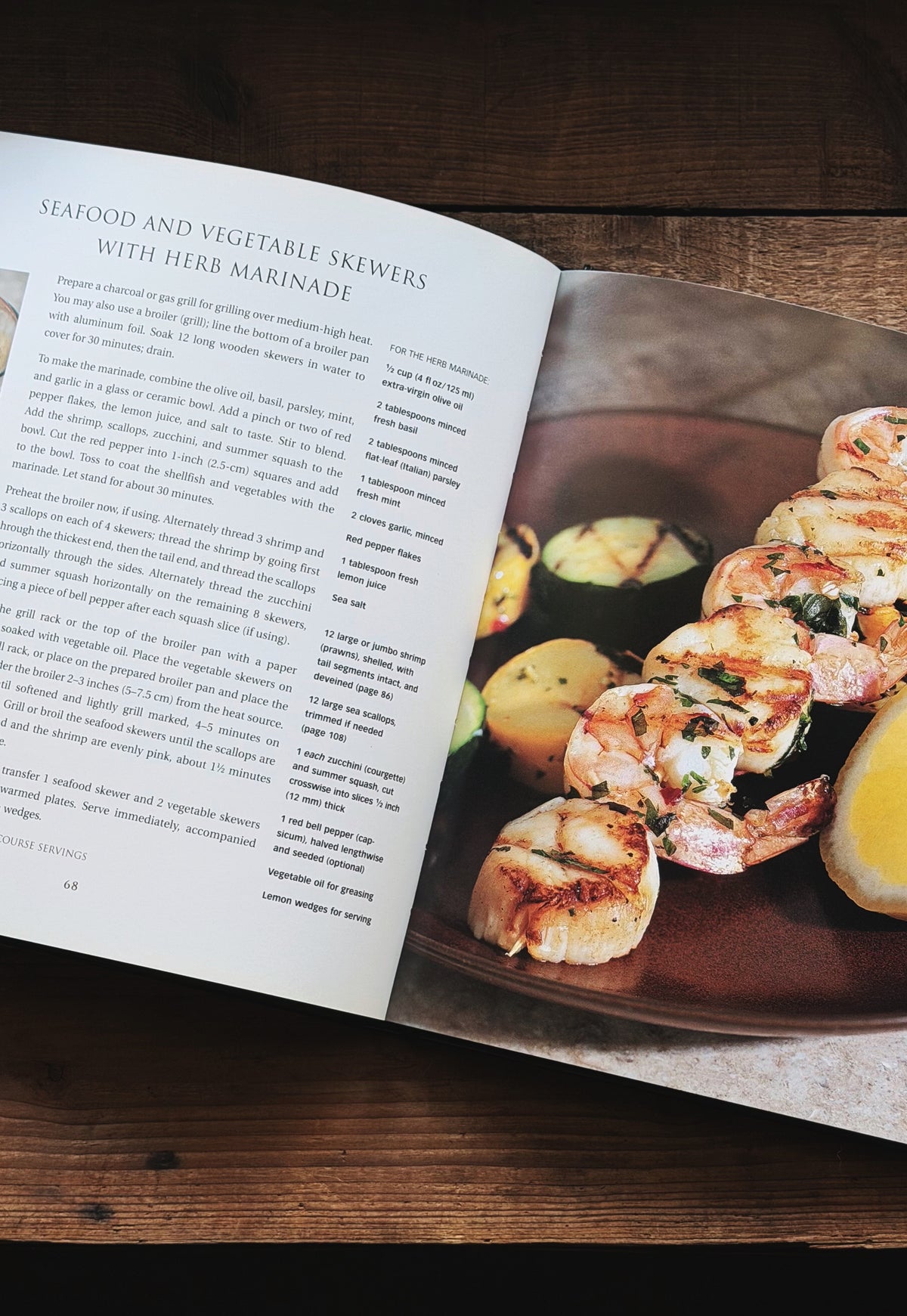 An open cookbook spread on a dark wooden table, showing a recipe page on the left and a plated seafood and vegetable skewer on the right.