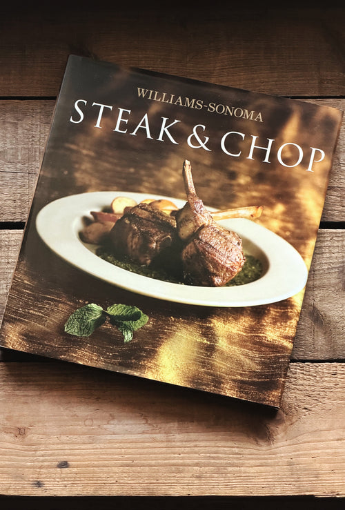 Williams-Sonoma Steak & Chop cookbook resting on a rustic wooden surface, angled slightly to the left. The book cover features a professional photograph of a cooked lamb chop on a white plate with herbs, set against a warm, golden-brown background with the title in white text.