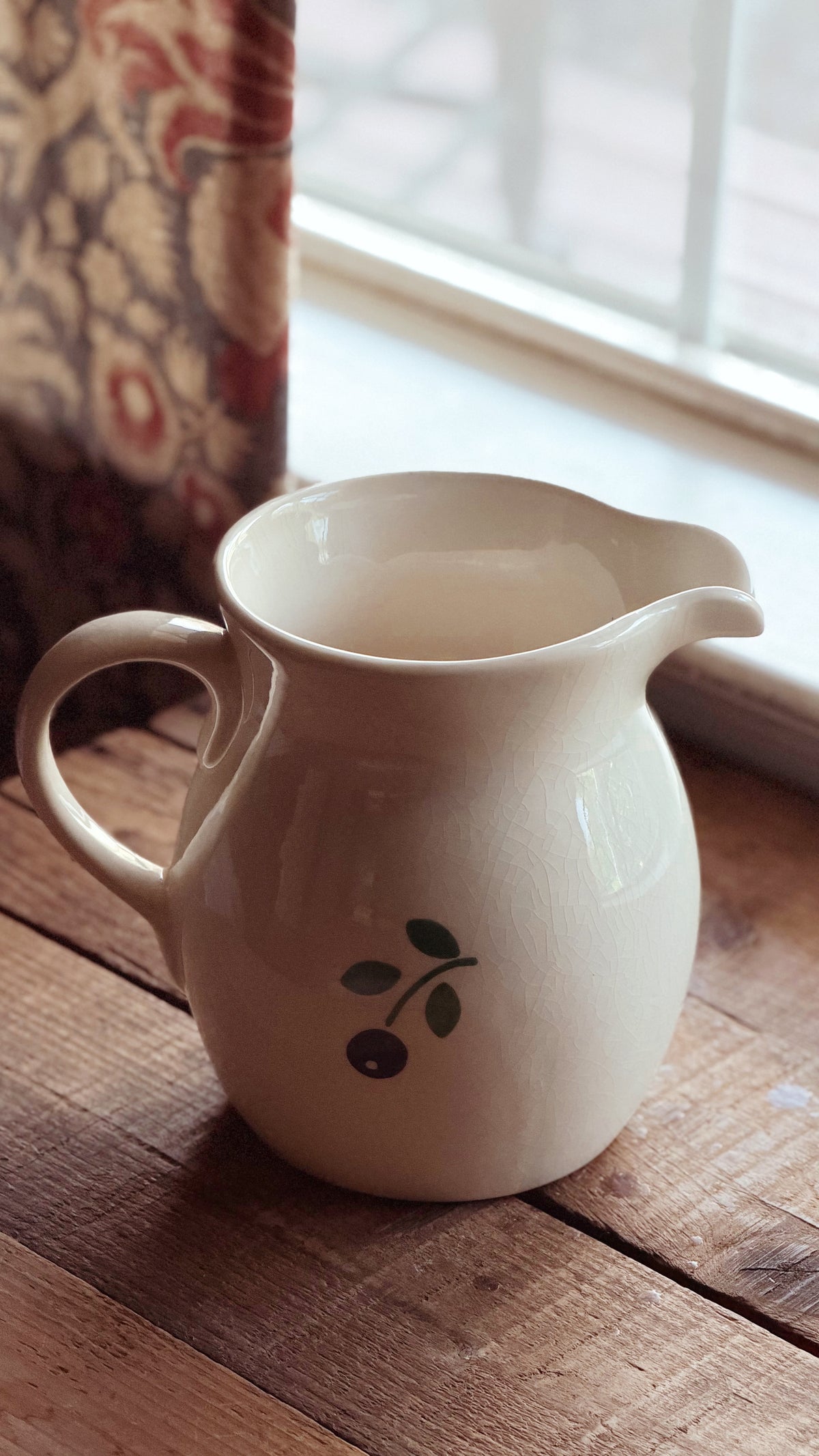 A cream-colored ceramic pitcher with a small green and black berry illustration, resting on a rustic wooden table near a window.