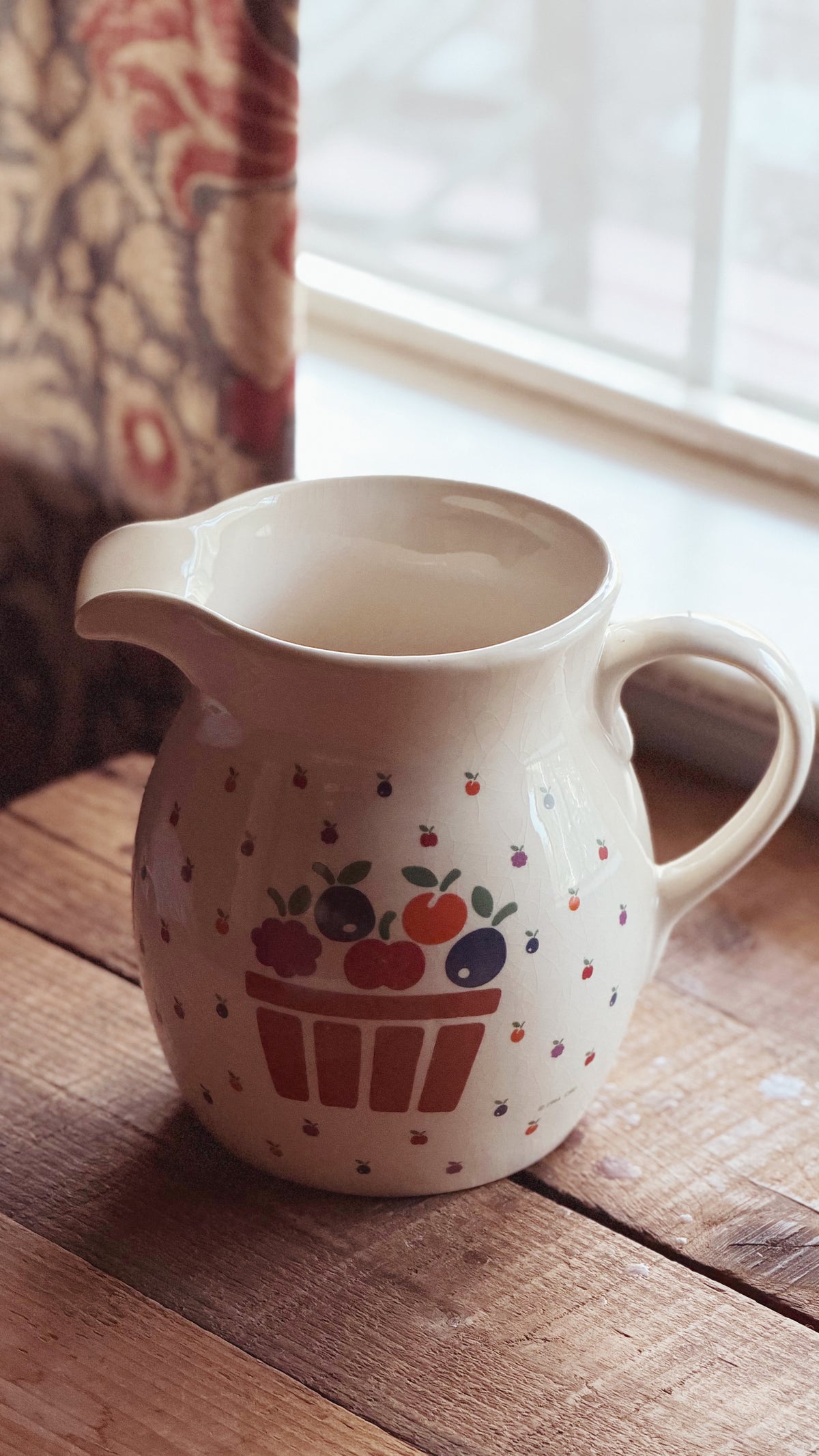 A cream-colored ceramic pitcher with a berry basket design, resting on a rustic wooden table near a window.