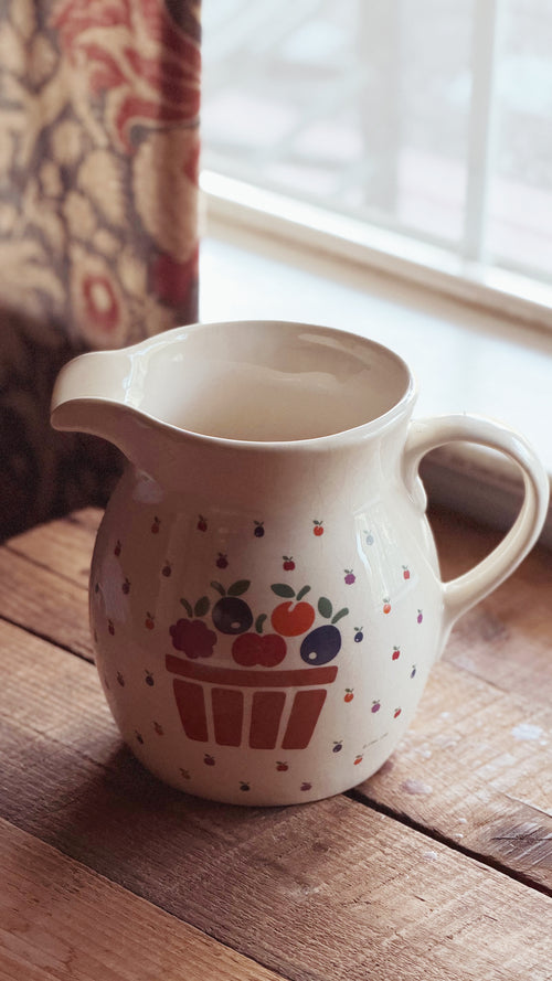 A cream-colored ceramic pitcher with a berry basket design, resting on a rustic wooden table near a window.