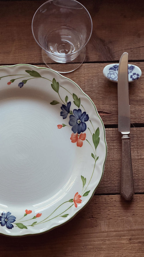 A white ceramic plate with a delicate floral border in blue, orange, and green, styled on a rustic wooden table alongside a clear glass and a knife.