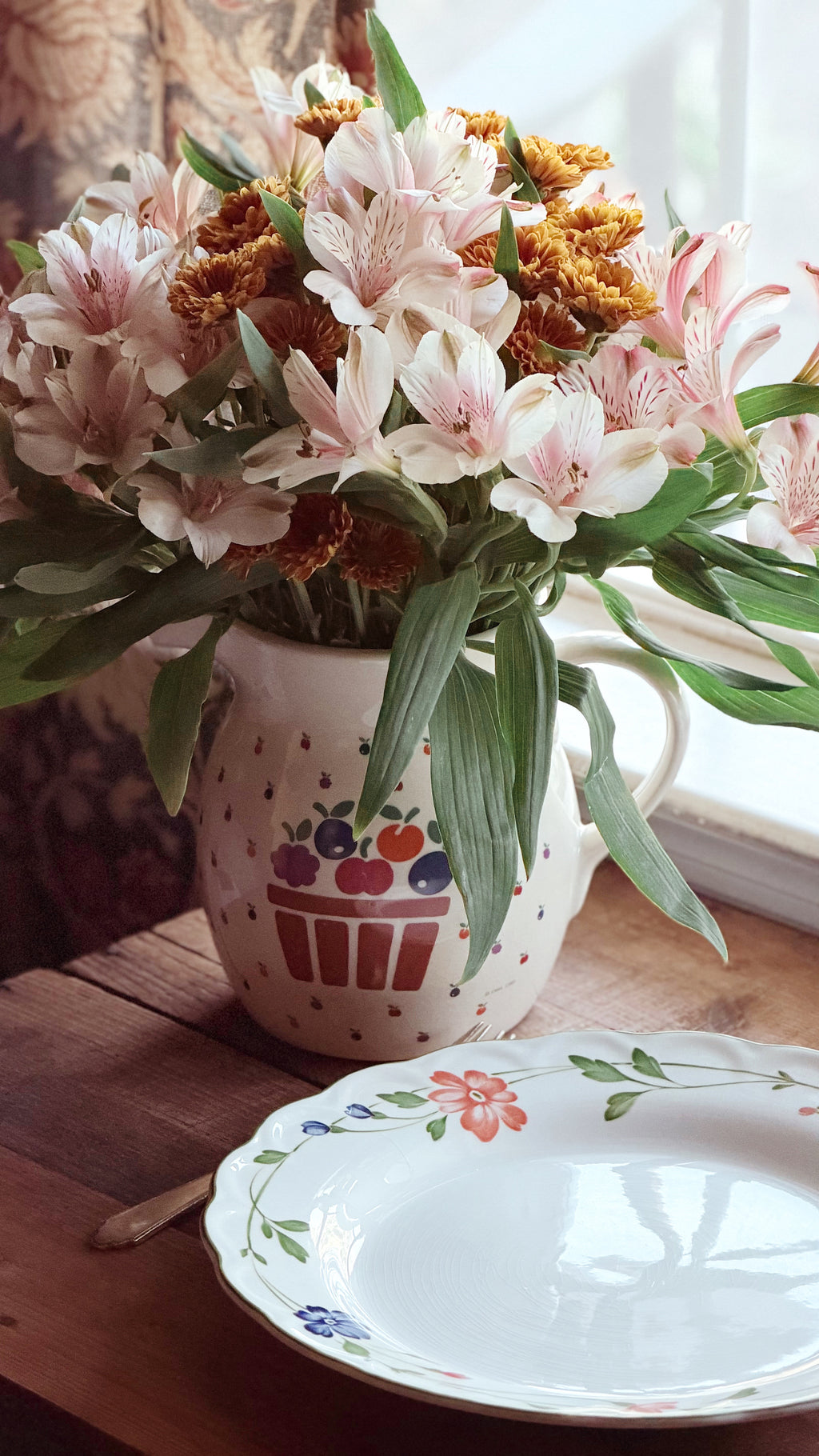 A ceramic pitcher filled with pink alstroemeria flowers and green leaves, placed on a wooden table next to a white floral plate in a softly lit indoor setting.