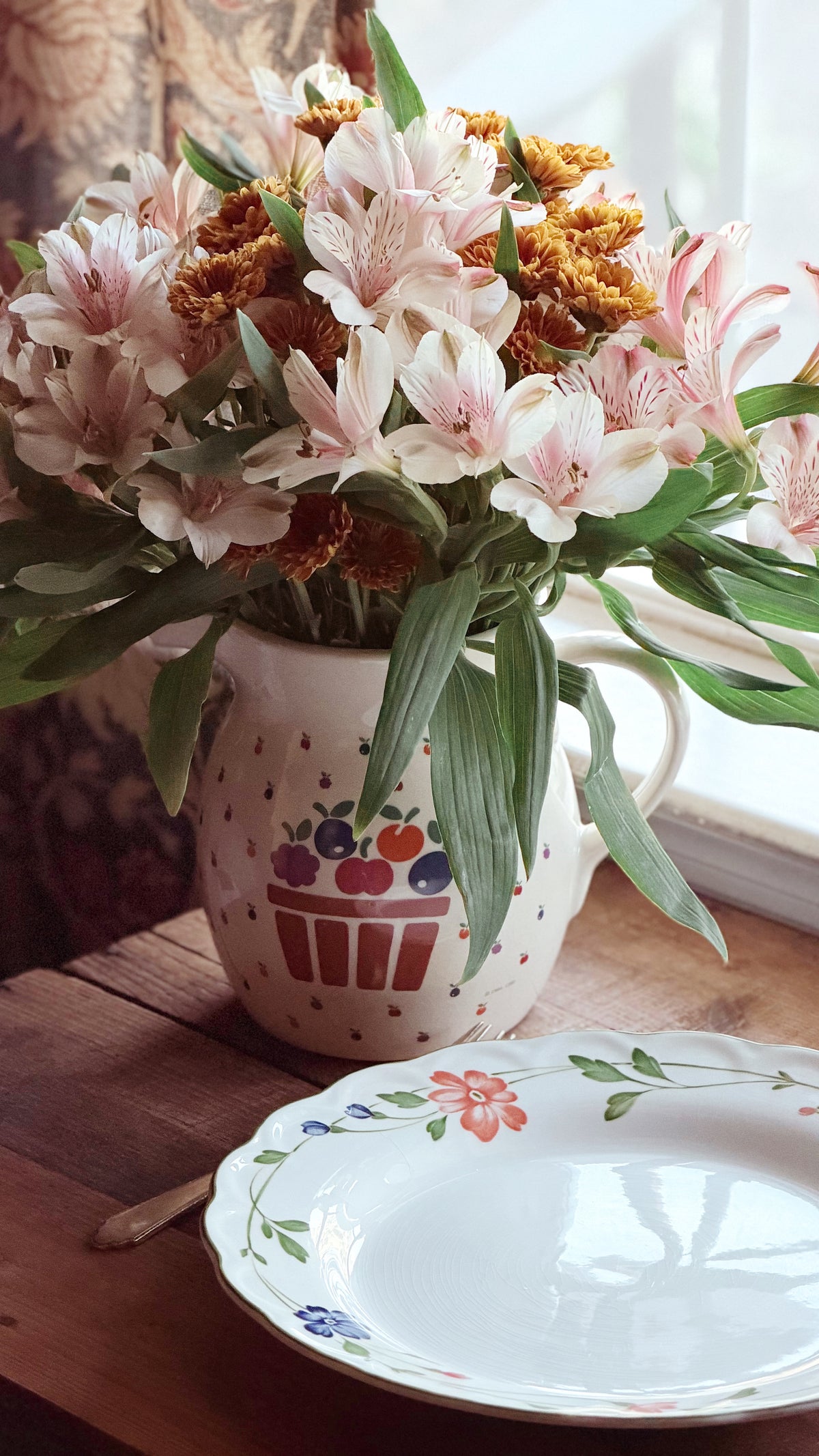 A ceramic pitcher filled with pink alstroemeria flowers and green leaves, placed on a wooden table next to a white floral plate in a softly lit indoor setting.
