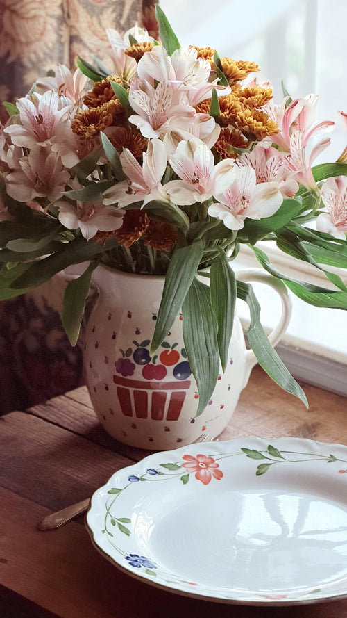 A ceramic pitcher filled with pink alstroemeria flowers and green leaves, placed on a wooden table next to a white floral plate in a softly lit indoor setting.