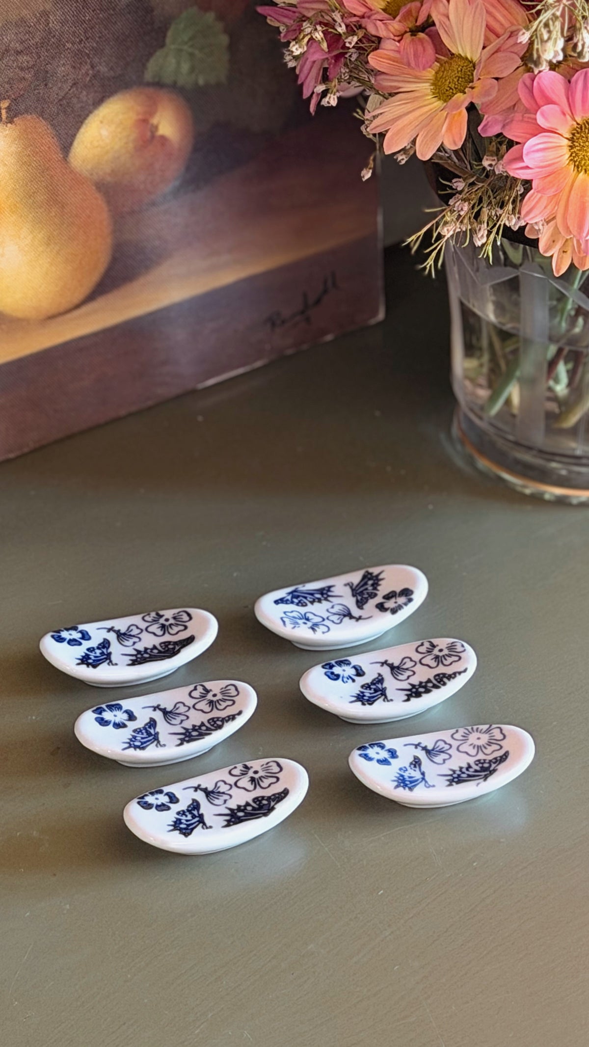 Five small white oval knife rests with blue floral patterns arranged on a tabletop, with a blurred vase of flowers and a fruit painting in the background.