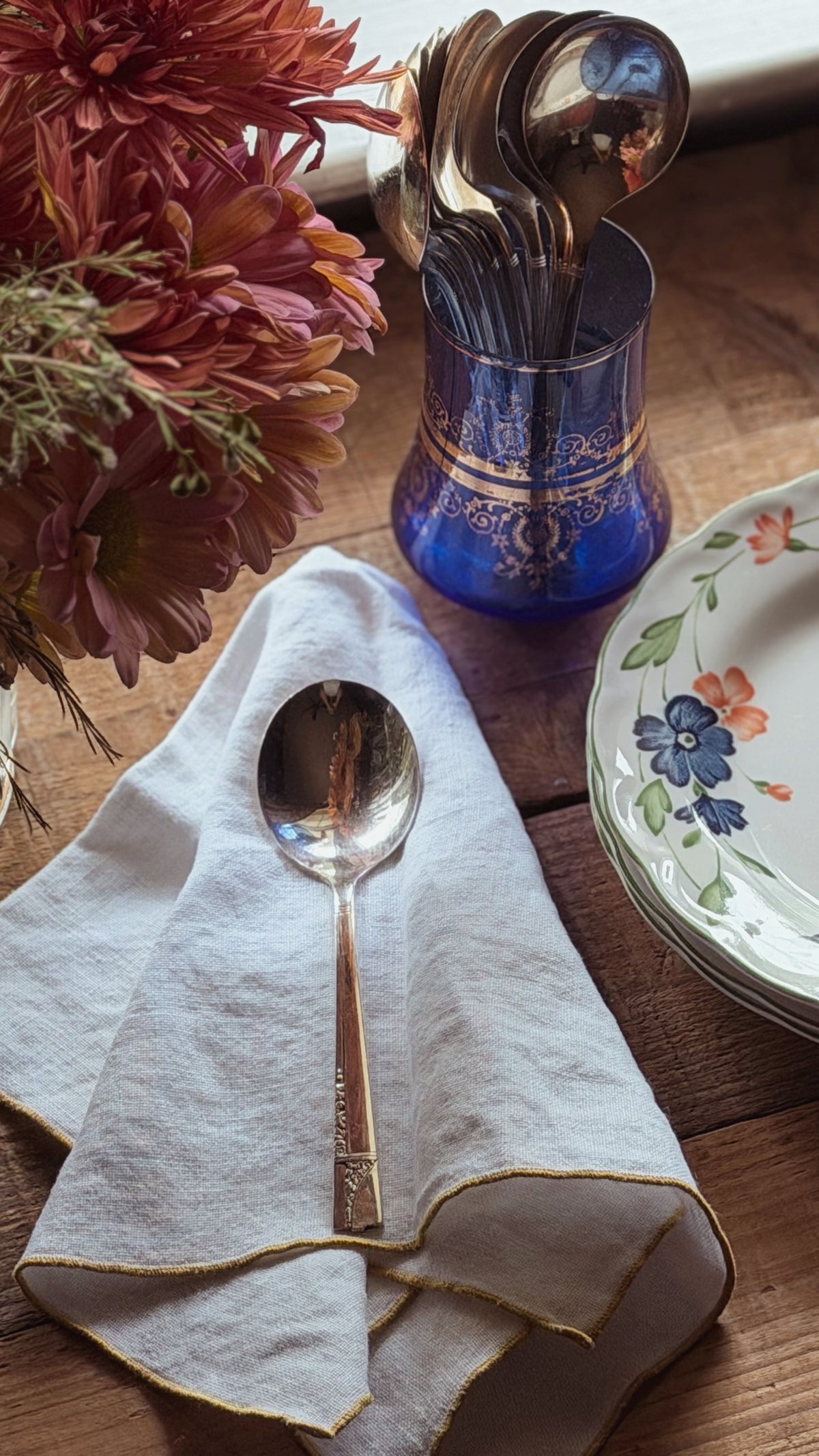 A silver metal spoon resting on a folded white linen napkin with gold trim, set on a wooden table next to a blue glass spoon holder and floral plates.