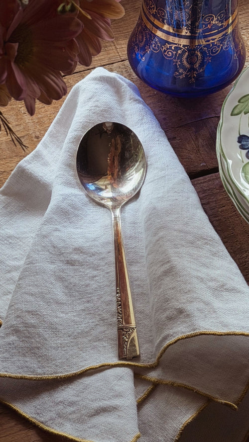 A vintage-style metal spoon with engraved detailing rests on a white linen napkin with gold trim, set on a rustic wooden table with a blue glass vase and plates in the background.