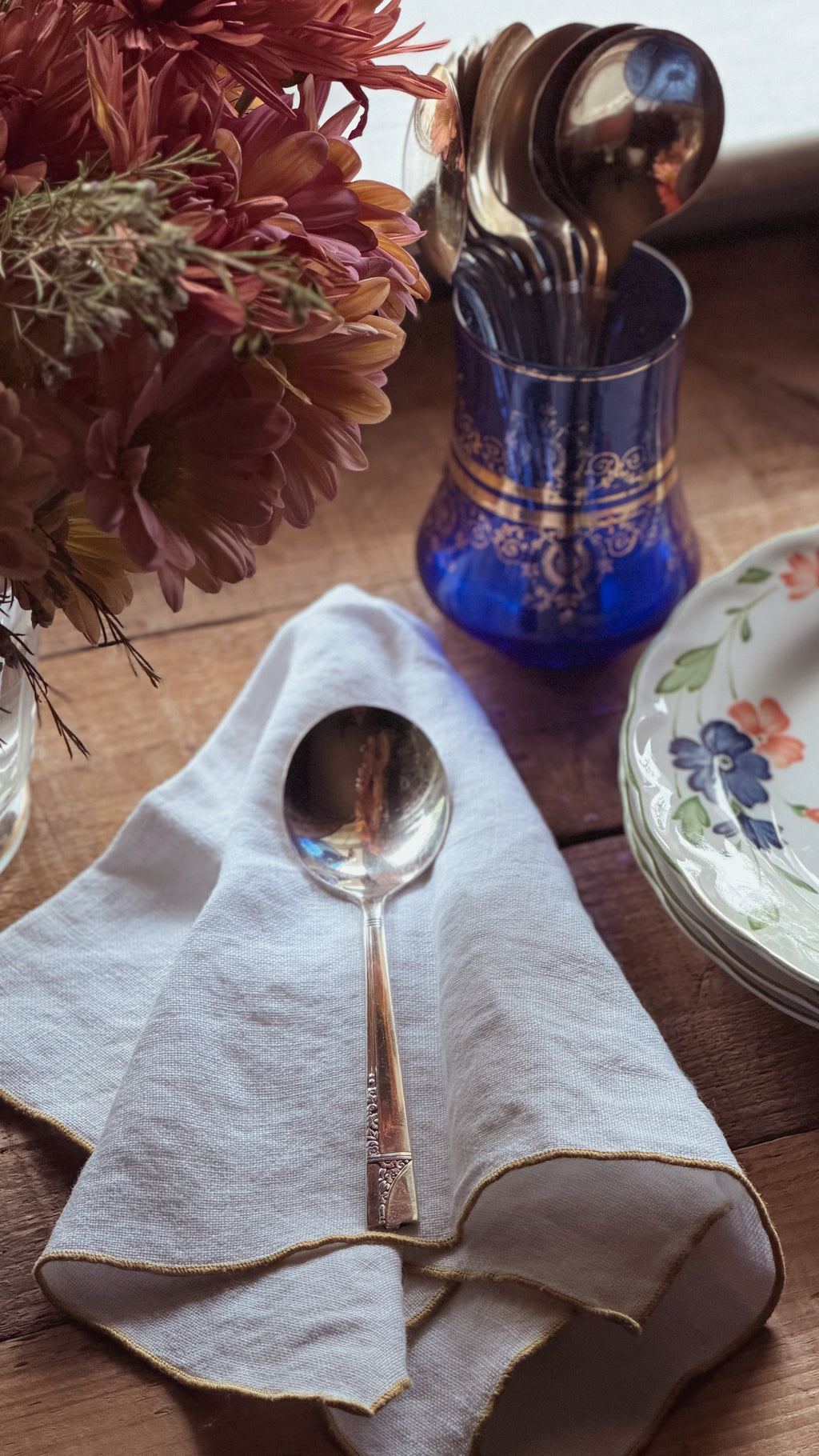 A polished silver spoon resting on a folded cream linen napkin on a wooden table, with a blue glass holder of cutlery, stacked floral plates, and dried flowers in the background.