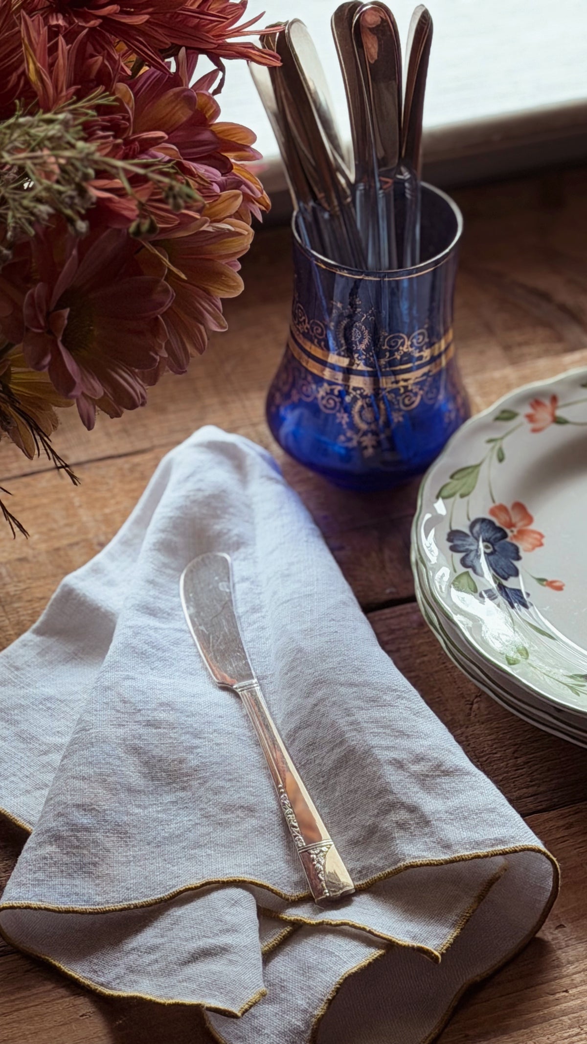 A styled tabletop scene with a folded white linen napkin holding a silver butter knife, a blue glass utensil holder with gold detailing containing metal cutlery, and part of a floral ceramic plate on a wooden surface.
