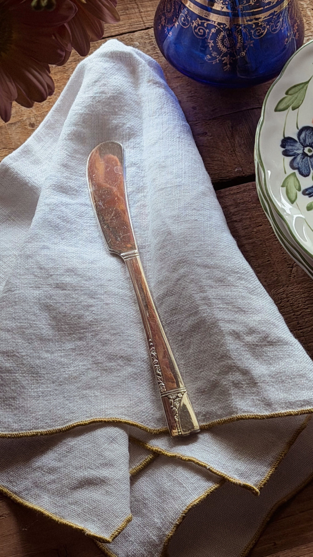 A vintage silver butter knife with a decorative handle, resting on a folded cream linen napkin with gold trim, set on a rustic wooden table with decorative plates and a blue glass vase in the background.