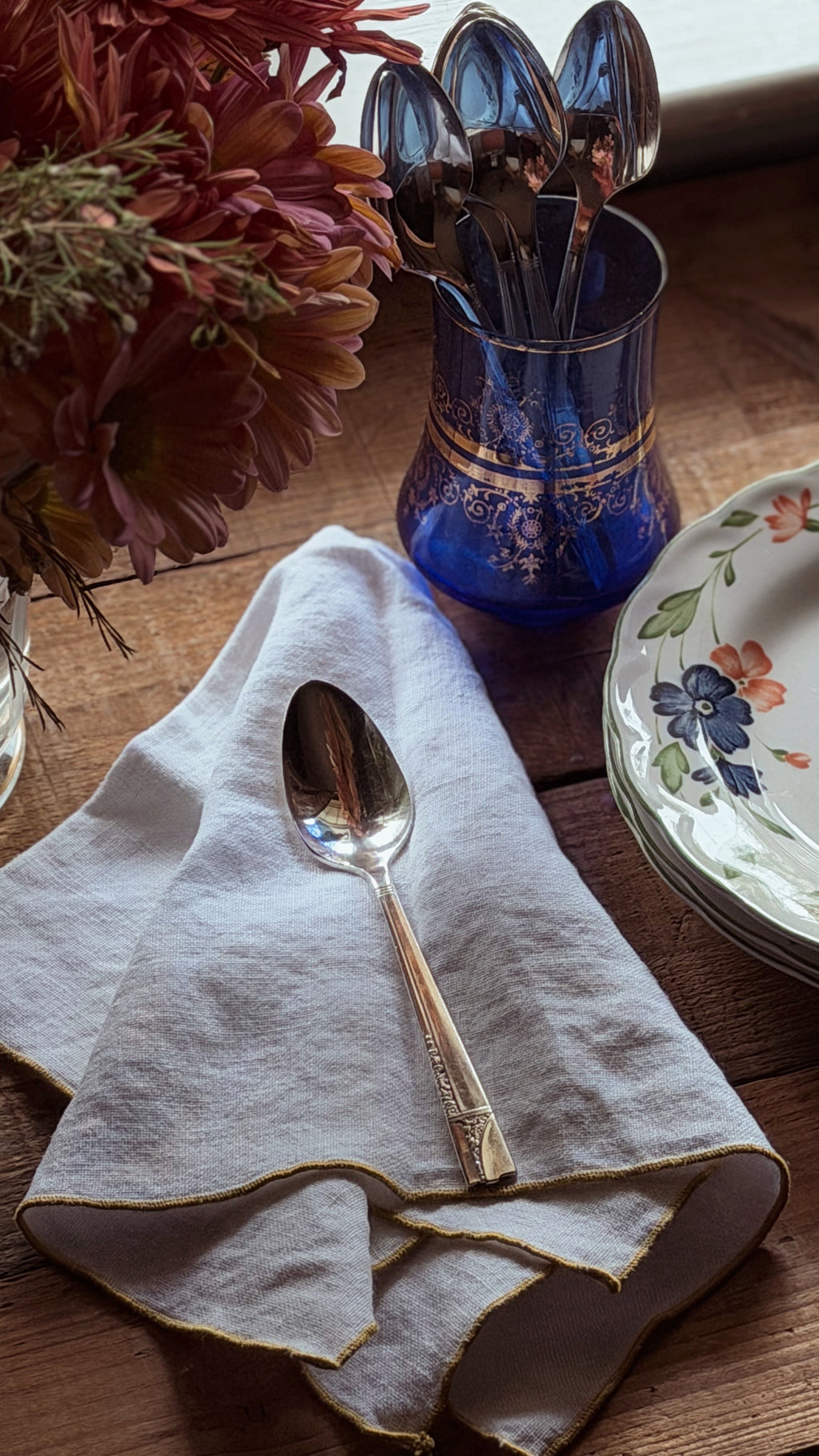 A folded light beige linen napkin with gold trim on a rustic wooden table, featuring a gold-handled spoon resting on it. In the background, a blue glass holder with gold detailing contains silver spoons, next to a floral-patterned plate and dried flowers.