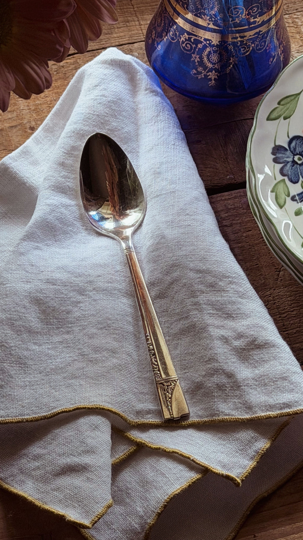 A vintage silver spoon with engraved detailing rests on a light beige linen napkin with gold-trimmed edges, set on a rustic wooden table alongside a blue glass bottle and a floral-patterned plate.