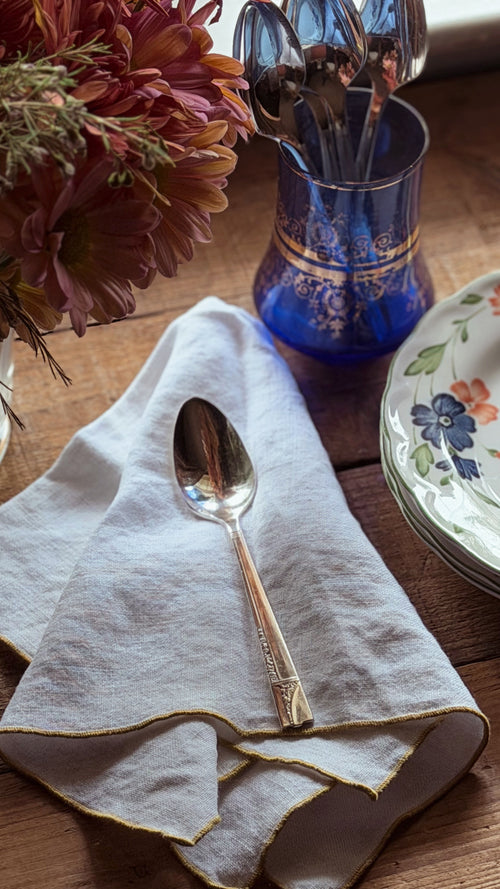 A close-up tabletop scene showing a folded white linen napkin with gold trim, a silver spoon resting on it, a blue glass utensil holder, and part of a floral plate on a wooden surface.