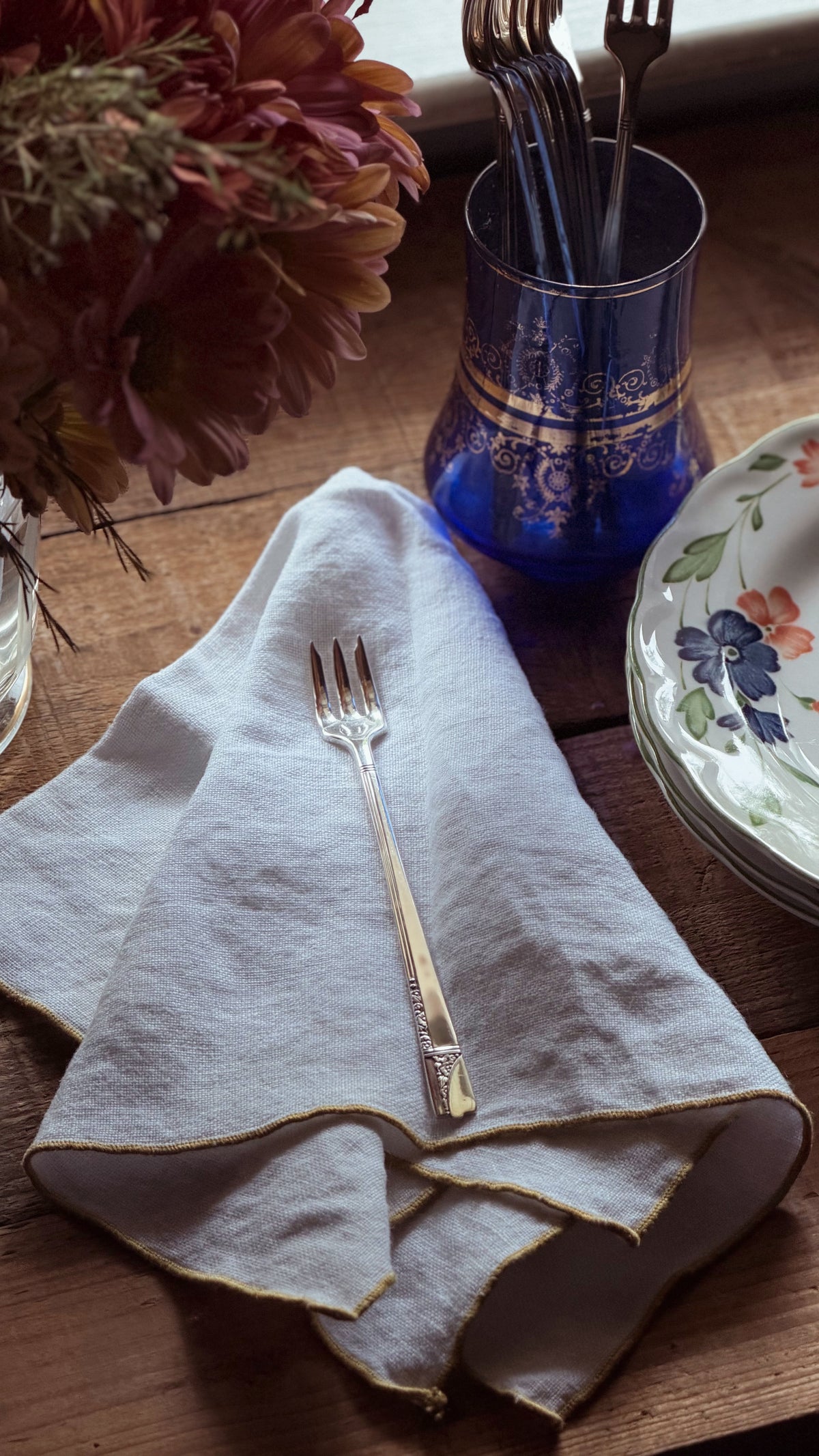 A folded white linen napkin with gold trim on a wooden table, featuring a silver fork on top, next to a blue glass cup holding forks and a floral plate.
