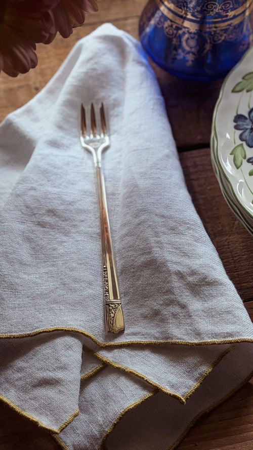 A vintage silver-toned metal seafood fork with engraved detailing, resting on a light beige linen napkin with gold trim, set on a rustic wooden table.