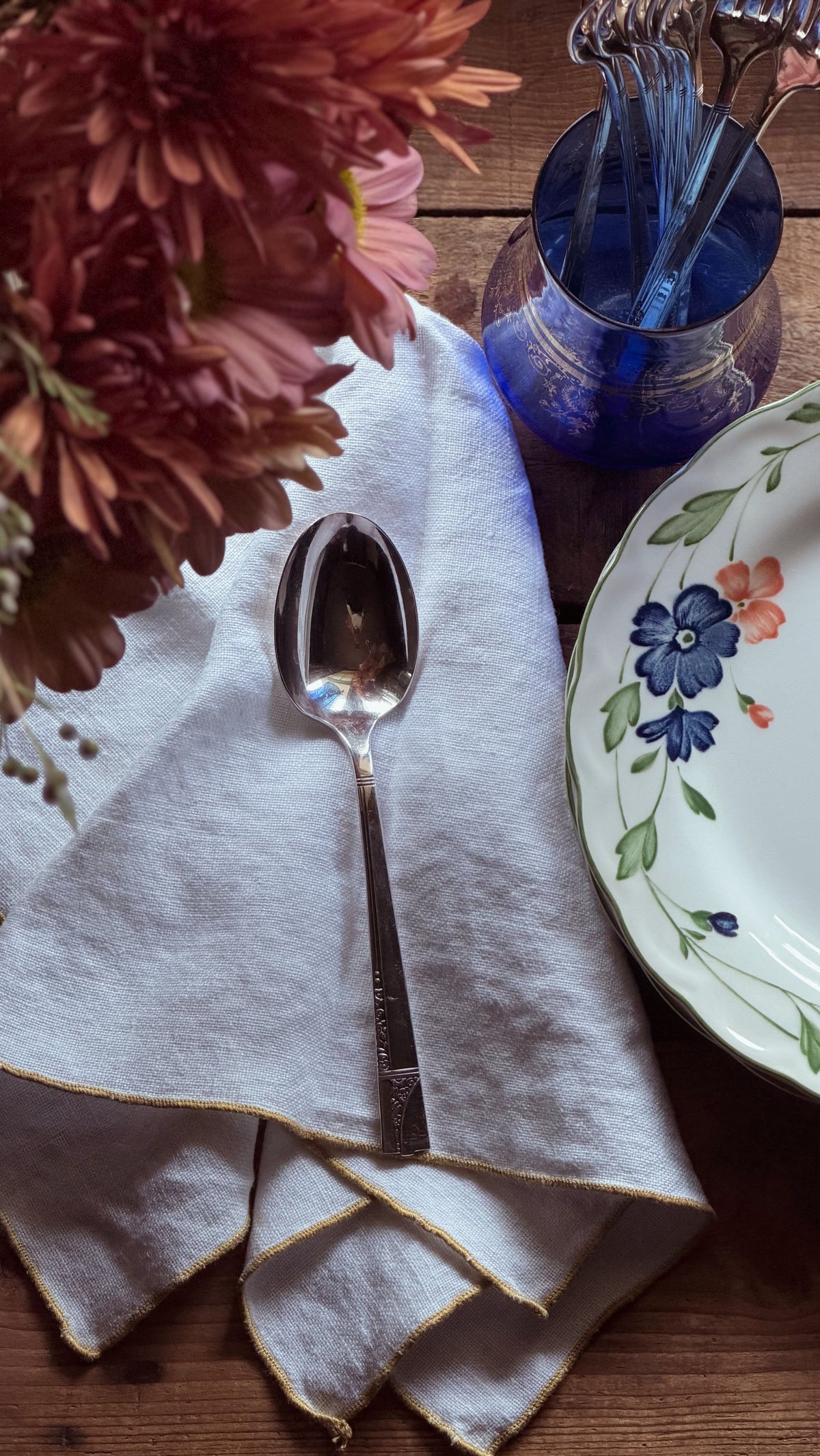A silver metal spoon resting on a folded white linen napkin with gold trim, styled on a wooden table alongside a blue glass cup, a floral-patterned plate, and flowers.