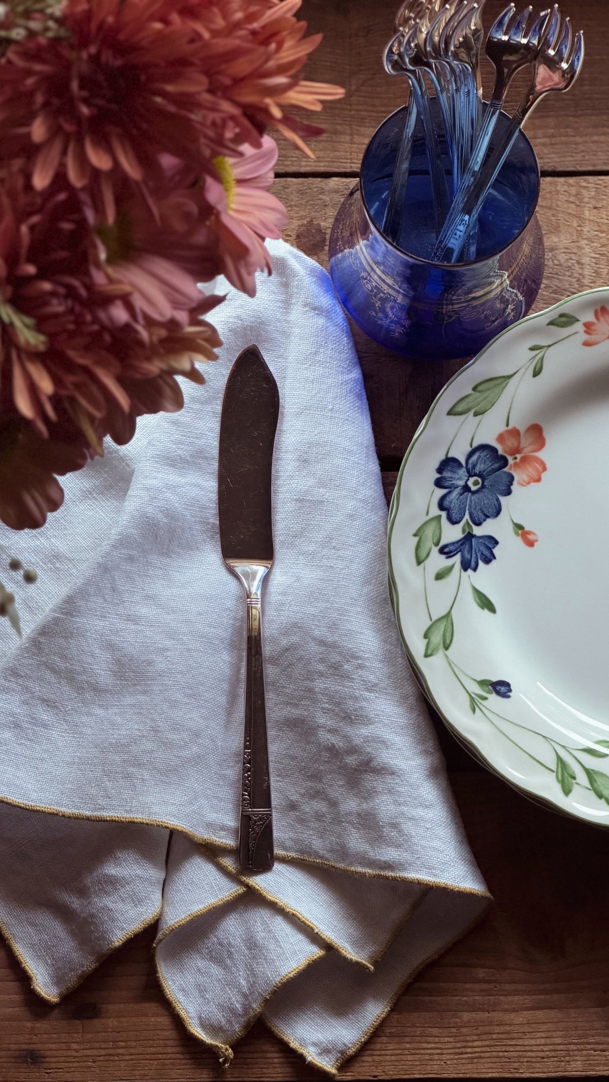 A styled tabletop scene with a folded white cloth napkin, a silver butter knife, a blue glass cup holding forks, and part of a floral ceramic plate on a wooden surface.