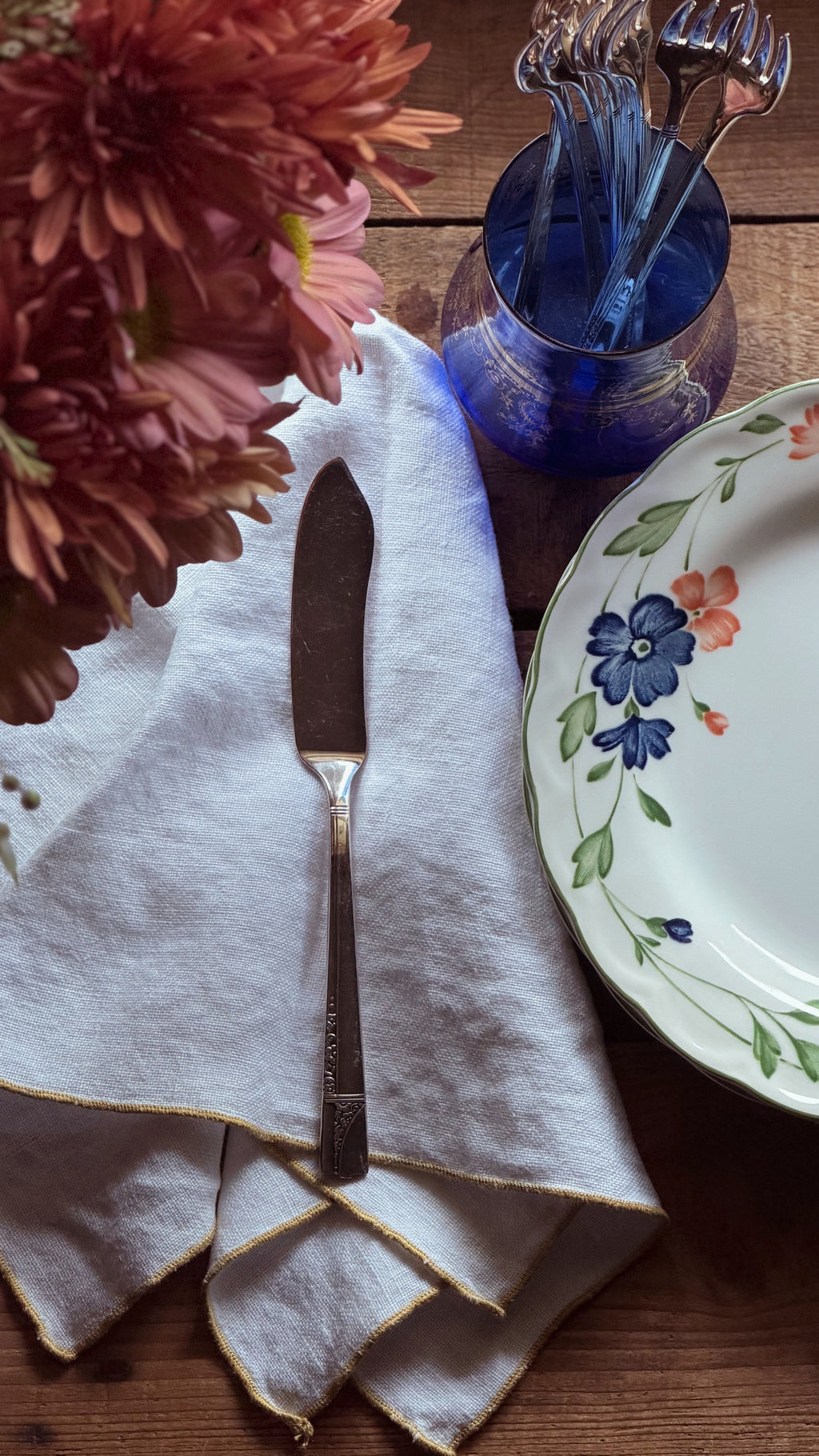 A styled tabletop scene with a white cloth napkin folded and laid on a wooden surface, a silver knife placed on top, a blue glass holder containing forks, and part of a floral ceramic plate visible at the right edge. Large pink flowers occupy the upper left corner.