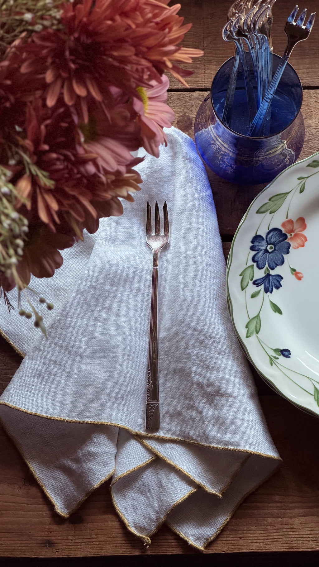 A folded white linen napkin with gold-trimmed edges on a rustic wooden table, styled with a silver fork, a blue glass cup holding forks, and a floral ceramic plate.
